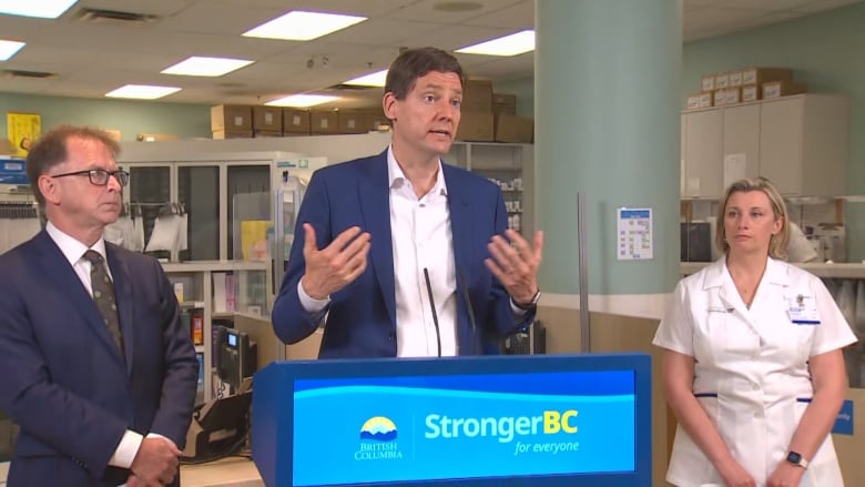 A man gestures off-camera as he answers questions at a news conference, flanked by another man in a suit, wearing glasses and looking serious with his arms folded. On the other side of the first man is a woman in a white smock. 