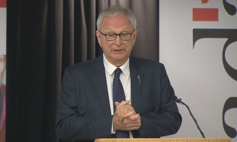 A man with grey hair and glasses wearing a suit and standing at a wooden podium.