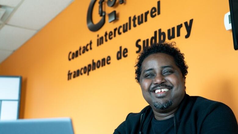 A smiling man standing in front of a yellow wall with black lettering that says Contact interculturel francophone de Sudbury.