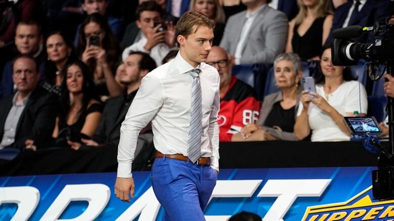 A man wearing a dress shirt and striped tie walks in front of a group of seated spectators.