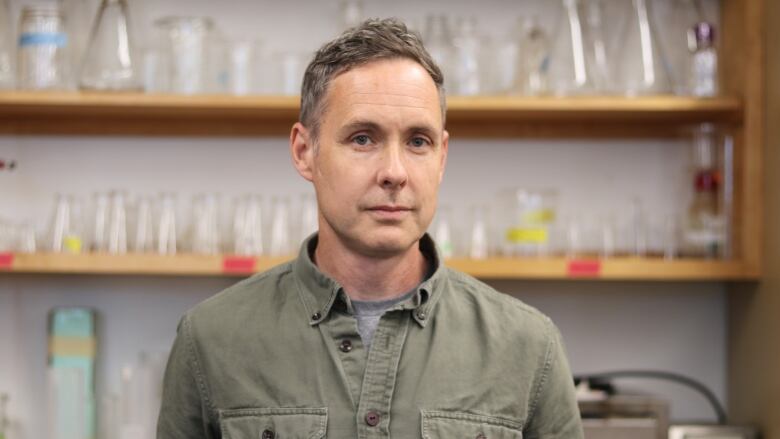 A man is wearing a collared, button up shirt. There are shelves of beakers and other scientific equipment behind him.