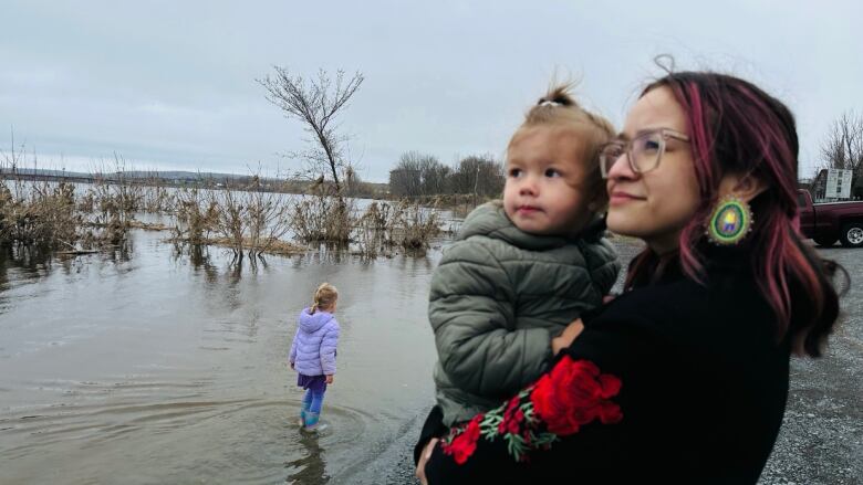 A woman holding a toddler looks out at a river. Behind her, another toddler plays in the water.