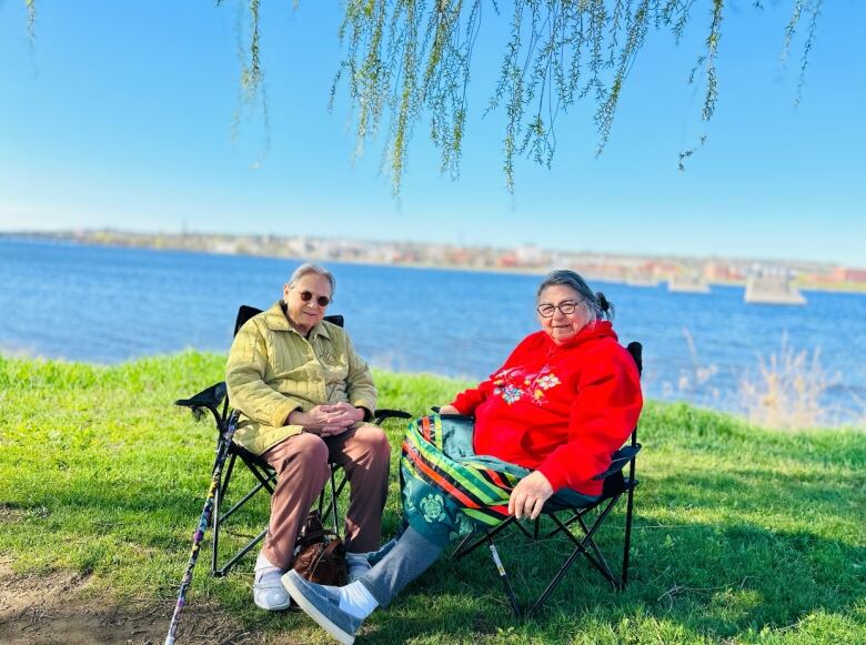 Two elderly women sit next to each other in lawns chairs. Behind them stretches a river.