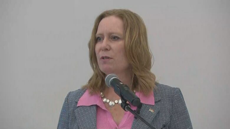 A woman in a pink blouse speaks into a microphone.