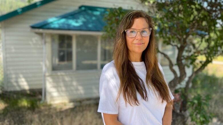 A young woman with long hair and glasses wearing a white blouse stands in front of a small white house with a green roof, smiling faintly.