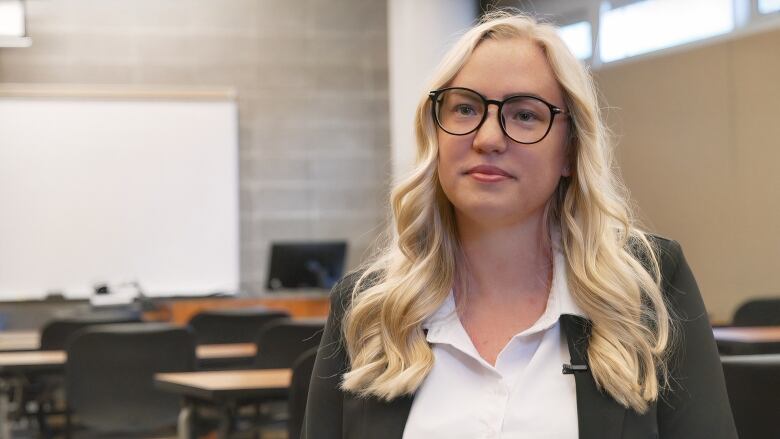 Close-up shot of a lady with blonde hair wearing black glasses, a white shirt and a dark green jacket.