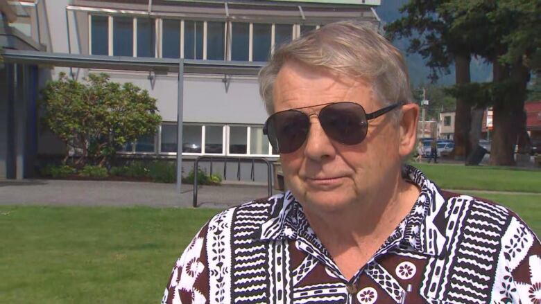 A man in a short-sleeved button down shirt wears sunglasses and stands in a yard speaking.