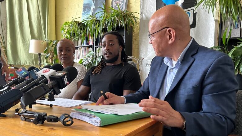 Three people sit behind a table in front of several media microphones. 