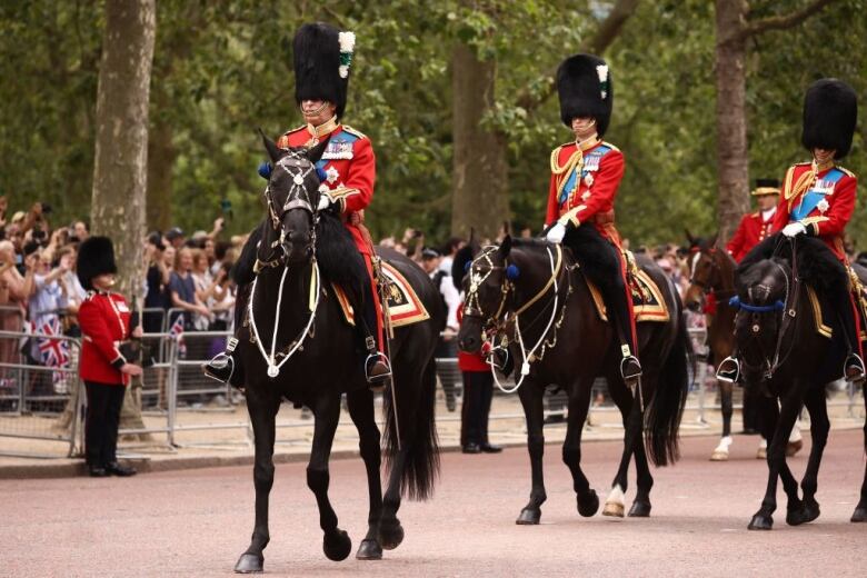 Three people wearing military uniforms ride horses in a parade with people watching from behind barricades.