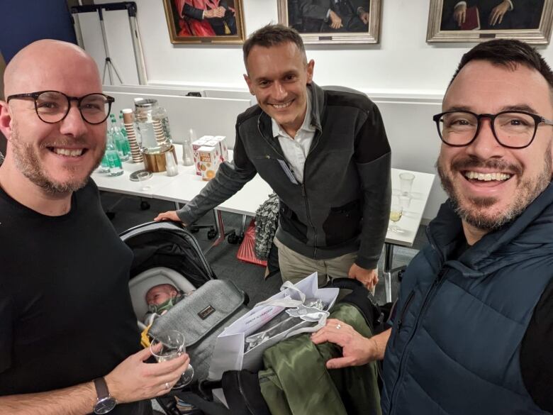 Three men gathered around a stroller carrying a sleeping baby, stand in front of a table with juice and cups and smile at the camera for a selfie.  