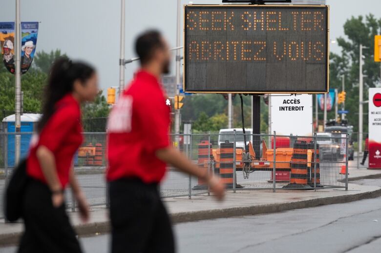 Two people wearing red pass in front of a sign that says 'Seek Shelter' in English and French.