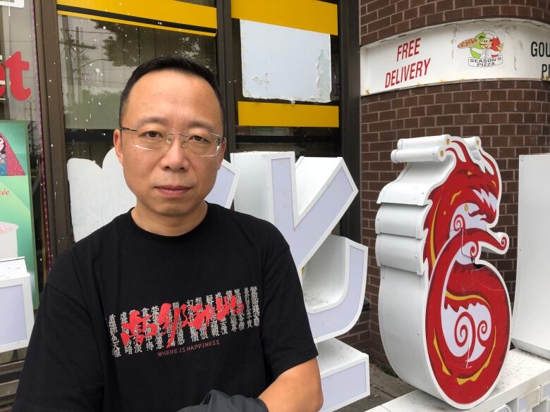 A Chinese man wearing glasses stares at the camera with a neutral expression wearing a black t-shirt. He is standing in front of the Chinatown logo and sign. 