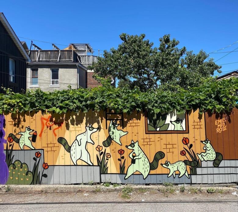 Daytime photo of a mural painted on a laneway fence. In a cartoon style, it depicts seven pastel raccoons. Some tag the wall with spraypaint. Others hang from fire escape ladders or hang out peacefully.