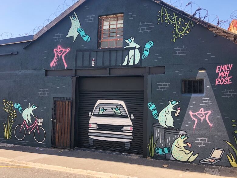 Mural on the exterior of a building. The walls are charcoal grey. Cartoon raccoons fill the space: riding bikes, driving a car, eating pizza, tagging the wall with spraypaint, scaling the roof.