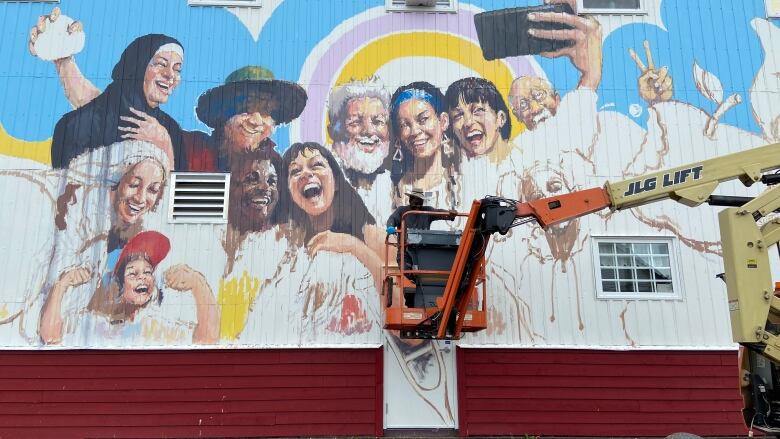 A man on a crane arm paints a large mural of many people smiling for a selfie on the side of a building.