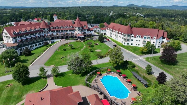 An aerial view of a long burgundy and white hotel. There is a bright blue pool, grassy areas and shrubbery in front of the main building.