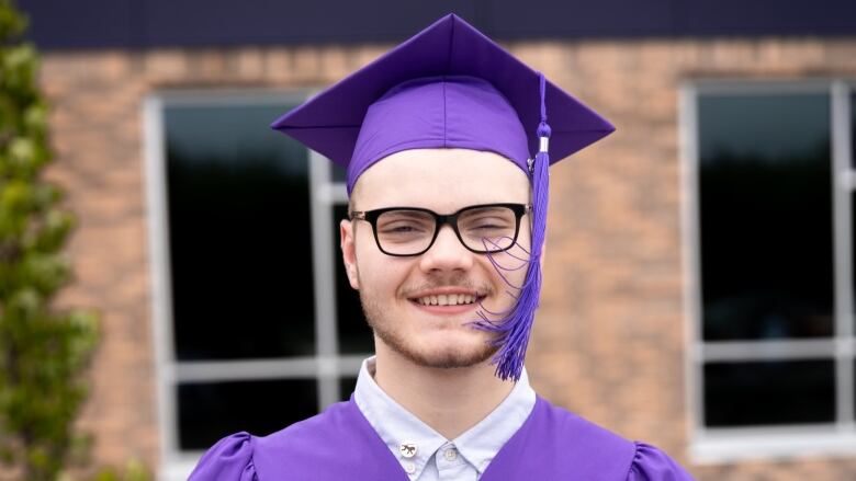 A man wears a purple graduation cap and gown. He holds a certificate of achievement with his name and the scholarship he won written on it.