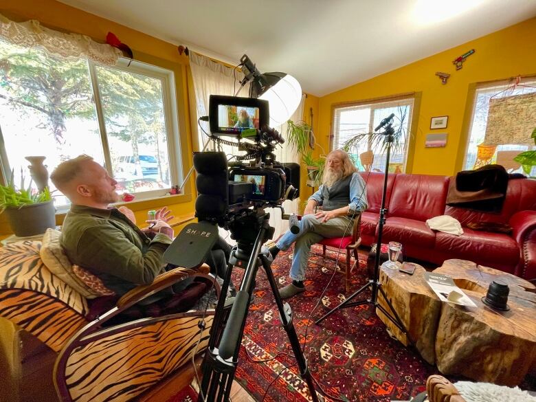 A man in profile sits on a chair behind a video camera. Another man with a beard and red hair sits in front of the camera on a sofa.