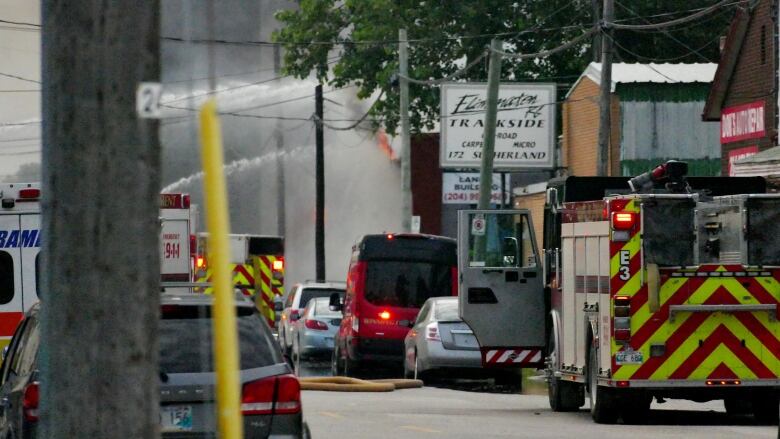 A fire hose sprays down a blaze on a street where business signs can be seen as well as parked cars among emergency vehicles.