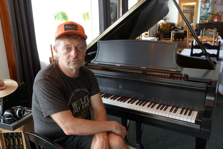 A man wearing a grey T-shirt and shorts, along with an orange baseball hat, sits at the bench of a piano in an otherwise empty dining room.