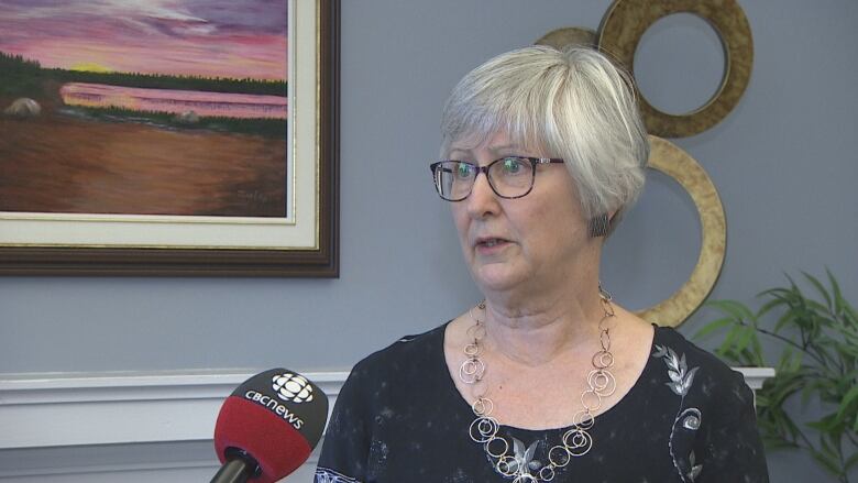 Grey-haired woman in glasses speaks into microphone. 