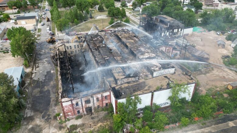 An aerial shot shows a large brick building with it's roof missing. Smoke is rising from the building and fire engines are spraying streams of water.