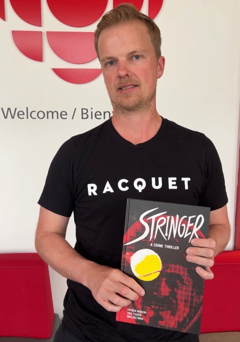 A man holds a copy of a hard cover graphic novel. The cover is black and red, and reads 