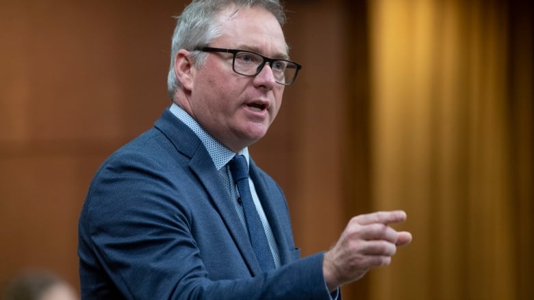 A man in glasses and a blue suit stands and speaks.