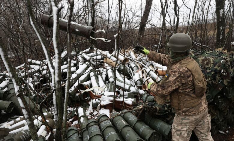 A soldier in camouflage military gear and a helmet tosses a large canister onto a large pile of similar canisters dusted with snow, piled in a leafless forest.