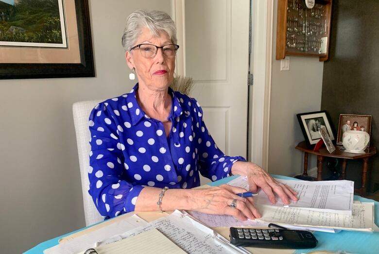 An elderly woman wearing a blue blouse sits at a kitchen table with papers in front of her.