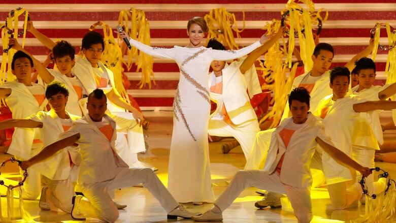 A woman wearing a white gown is surrounded by male dancers as she performs onstage.