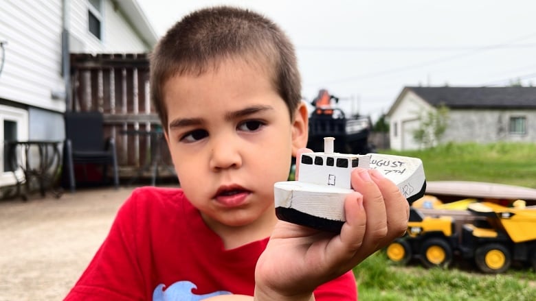 A little boy in a red shirt holds up a small white boat 