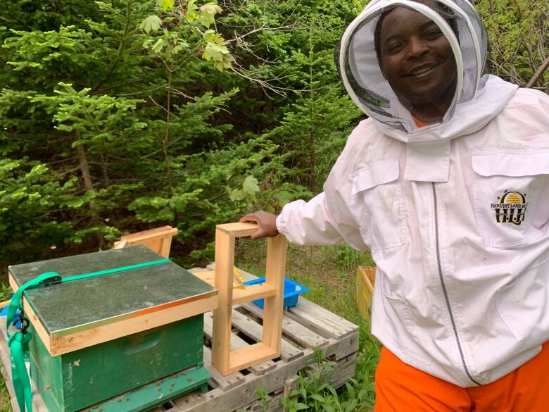 A smiling man wearing a beekeeping suit stands in front of a hive box.