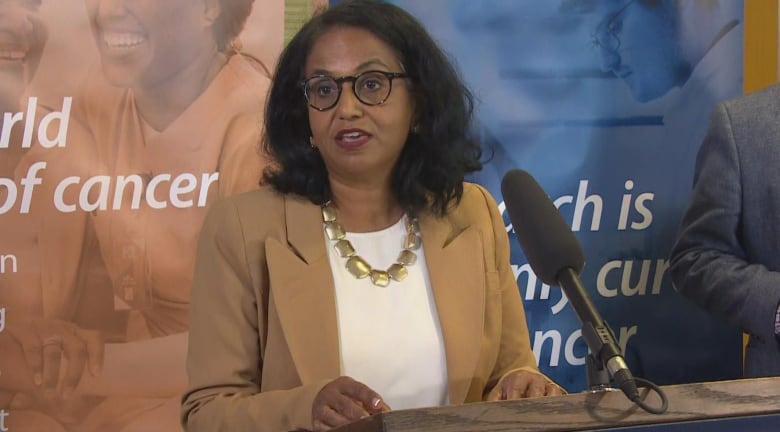 A woman wearing black rimmed glasses and a pink blazer is shown speaking at a press conference.