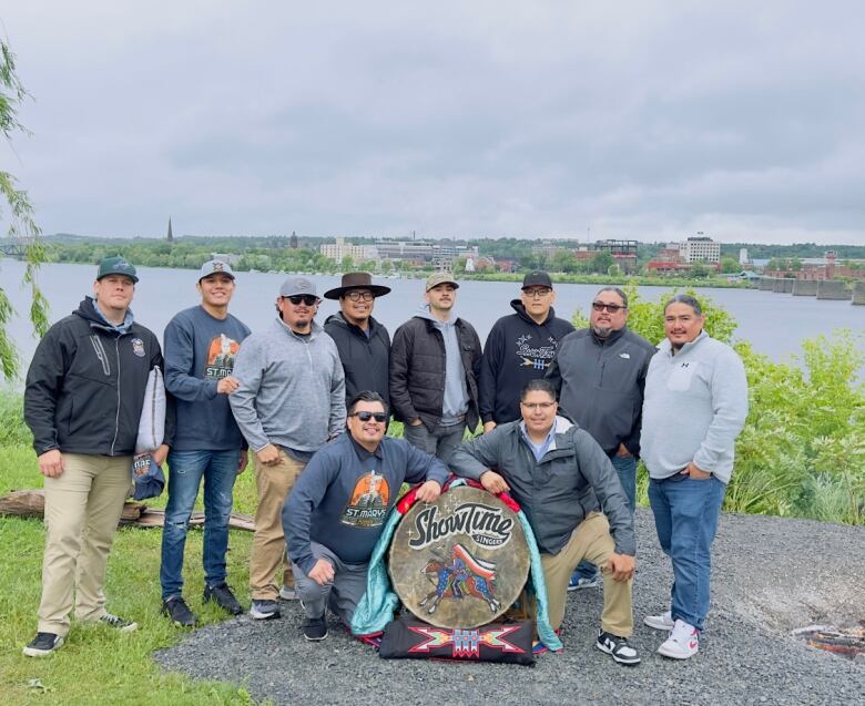 A group of men stand together in front of a river. In front of them sits a drum.