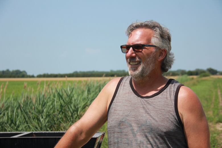 A farmer wearing a tank top stands in his field smiling off camera.