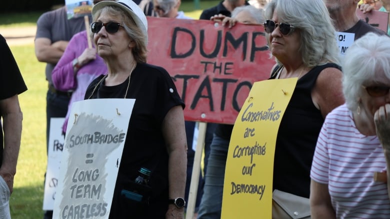 Two protesters stand outside on a sunny day with signs around their neck. One sign reads, 