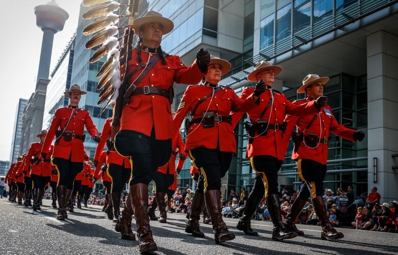 members of the RCMP in their fancy red dress uniforms march in a parade formation on a downtown Calgary street with the Calgary tower visible behind them.