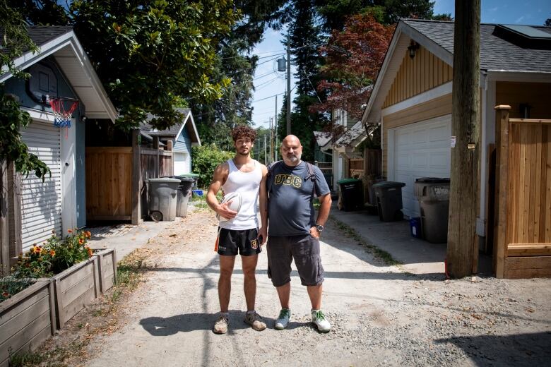 A young man in his late teens and a man in his fifties stand arm-in-arm in a residential laneway