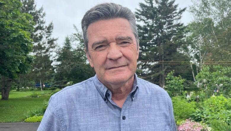 A man with greying hair, wearing a blue, short-sleeve collared shirt, smiles faintly, surrounded by trees and plants.