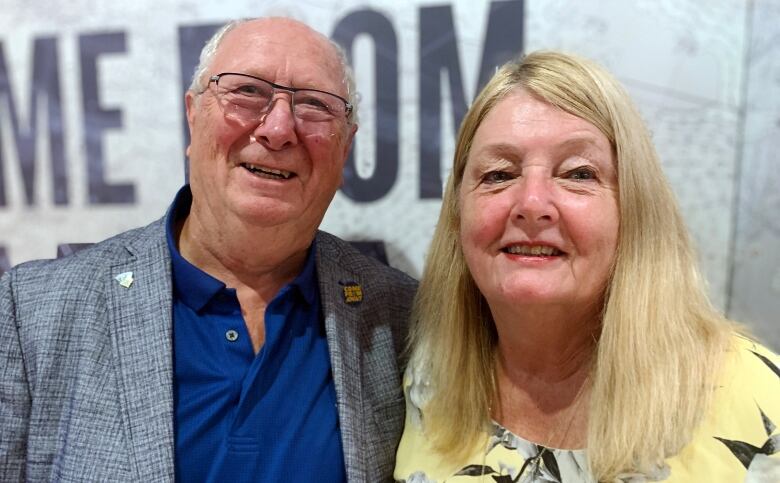 A man with glasses and a woman with blonde hair smile in front of a Come From Away sign.