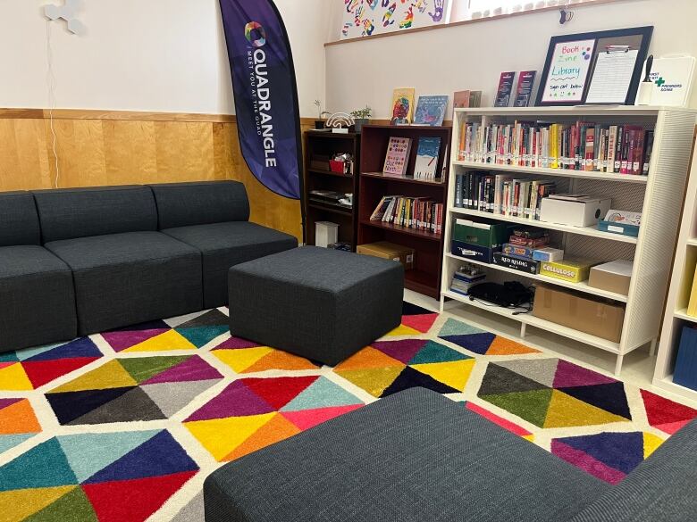 A colourful carpet sits under a grey sectional sofa, with three bookshelves holding novels, paintings, and supplies to the right. A Quadrangle banner hangs in the corner. 