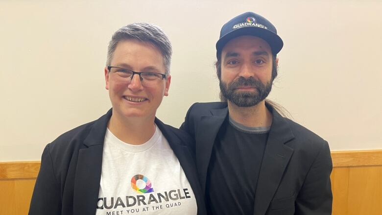 Two people, one in a black shirt and one in a white shirt, stand side by side in front of a beige wall. 