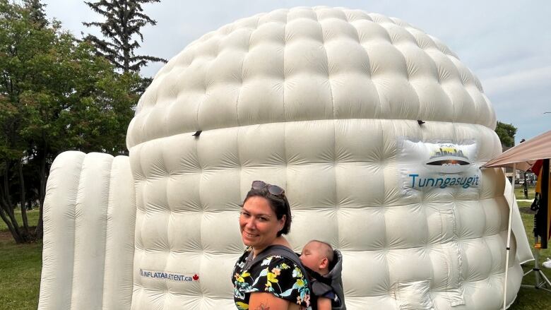 A woman smiles to the camera. She's carrying a sleeping baby on her back, and standing in front of an inflatable igloo.