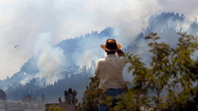 A man wearing a beige shirt, blue jeans and tan cowboy hat watches as a wildfire burns on a hillside in the distance.