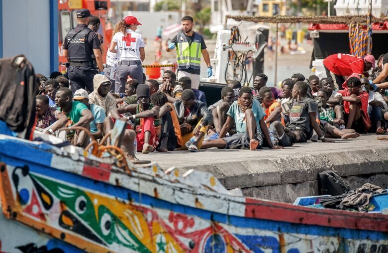 People rest on a pier while rescue workers are seen behind them and a boat is seen in the foreground.