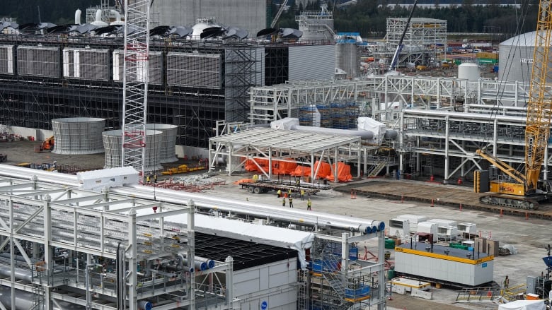 A large factory facility is seen with workers off in the distance. Cooling towers and cranes are visible.