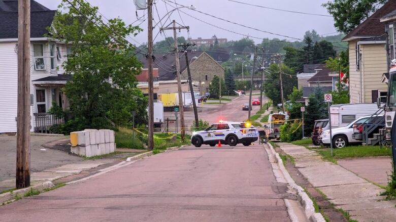 A residential street with several homes, driveways and signs with a police SUV and crime scene tape.