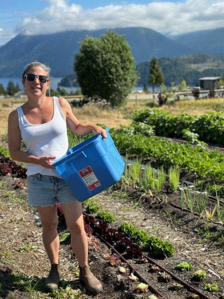 A woman is pictured standing in a field of vegetable crops, mountains and blue sky in the background. 