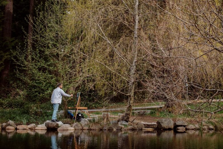 An artist uses an easel and palette to paint next to a scenic lake.